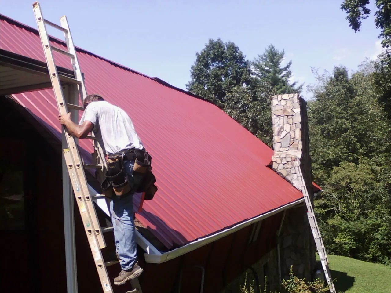 Roofer on a ladder beside a red metal roof and stone chimney in Dunsmuir